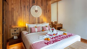 Quarto de hotel com teto de madeira, cama com roupa de cama branca, almofadas rosa e brancas, um painel de parede redondo feito de material trançado e uma mesa de cabeceira com um telefone.