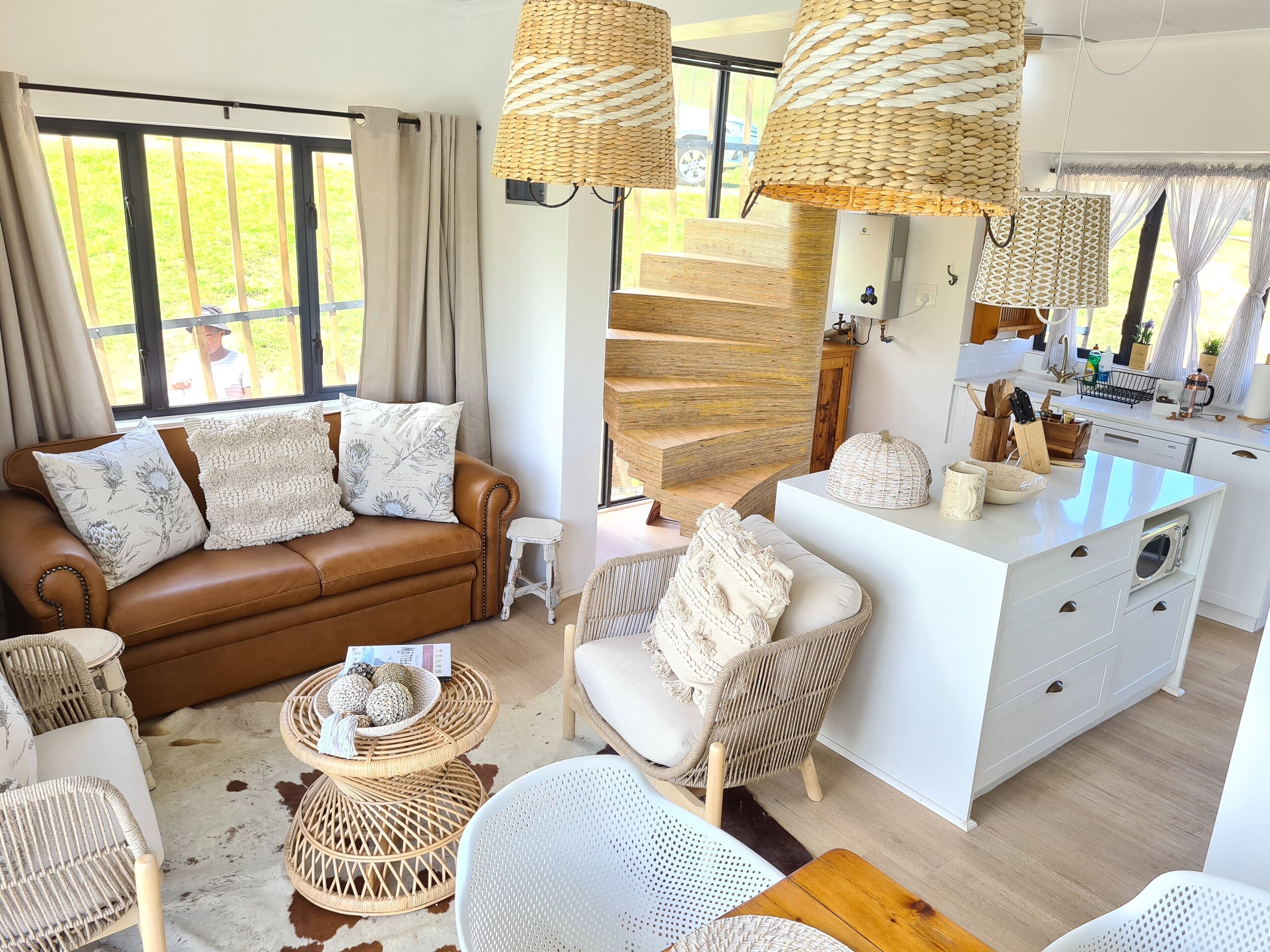 A modern living room with a brown leather sofa, white chairs, wicker side tables, and a kitchen area with white cabinets and a wooden island.