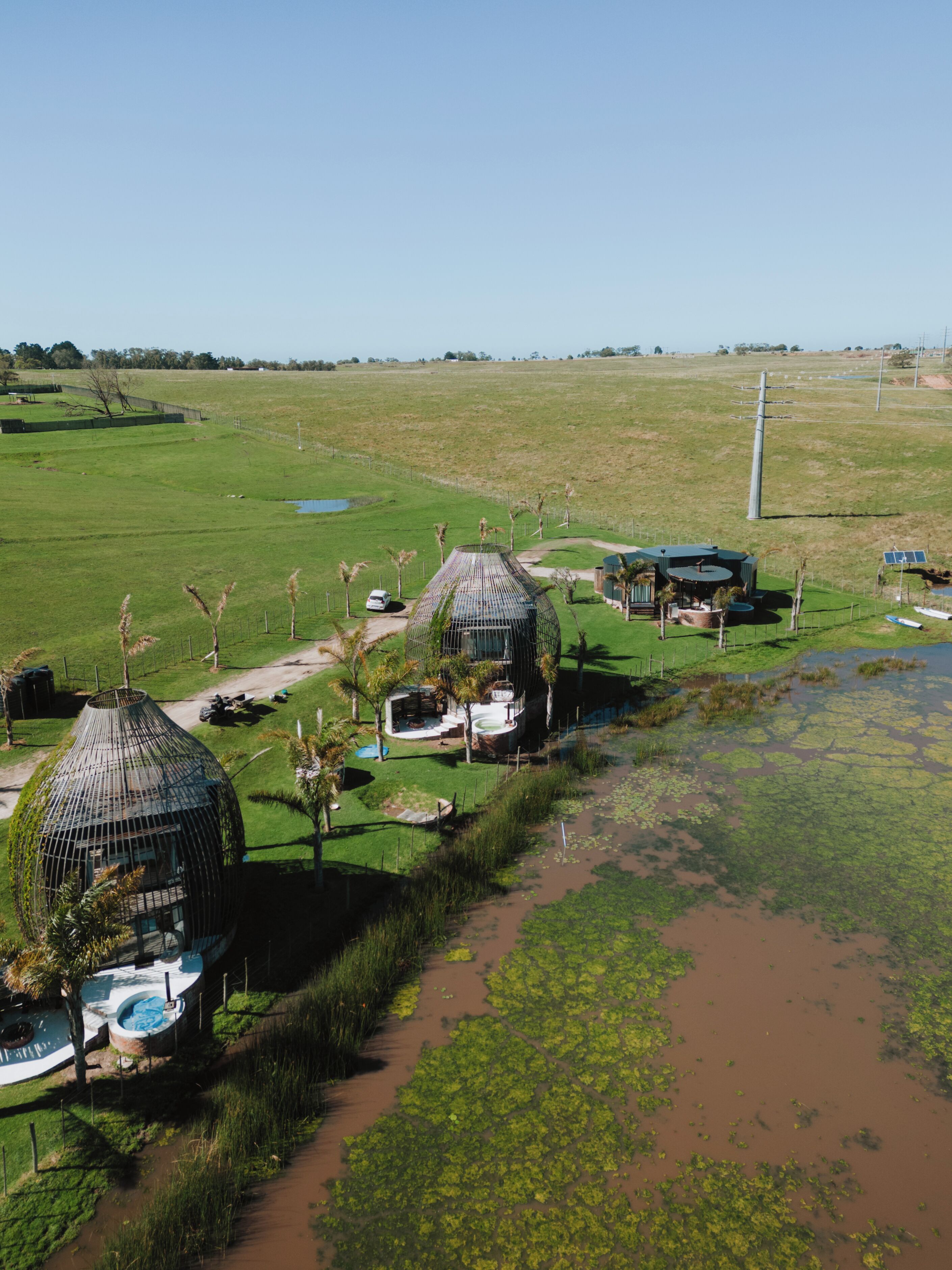 Ländliche Szenerie mit zwei kuppelförmigen Gebäuden, vermutlich Gästeunterkünfte, umgeben von üppiger Vegetation und einem kleinen Gewässer.