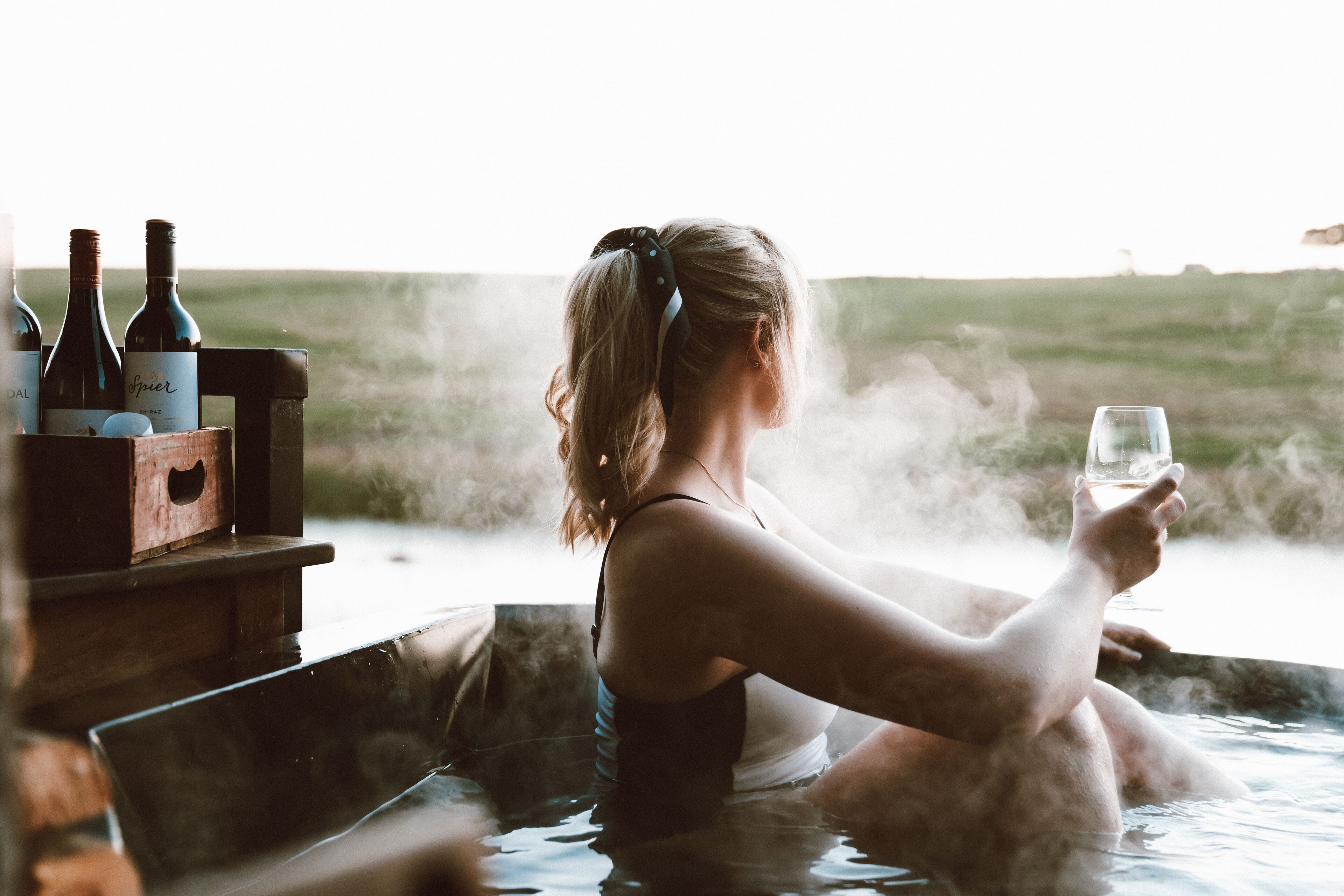 Una persona relajándose en un jacuzzi con botellas de vino y una copa.