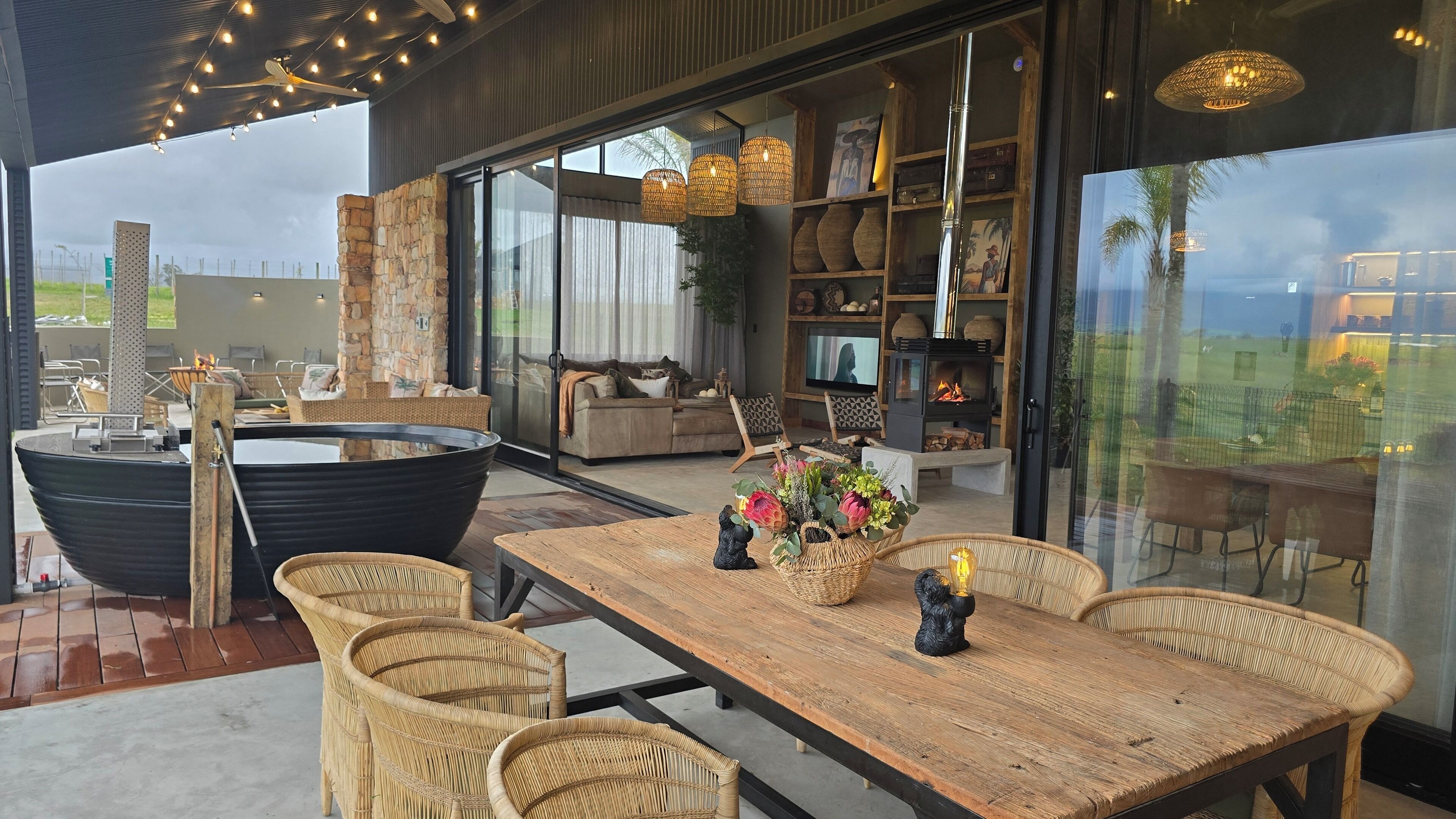 A modern living room with a large wooden table, wicker chairs, a stone wall, and a fireplace.