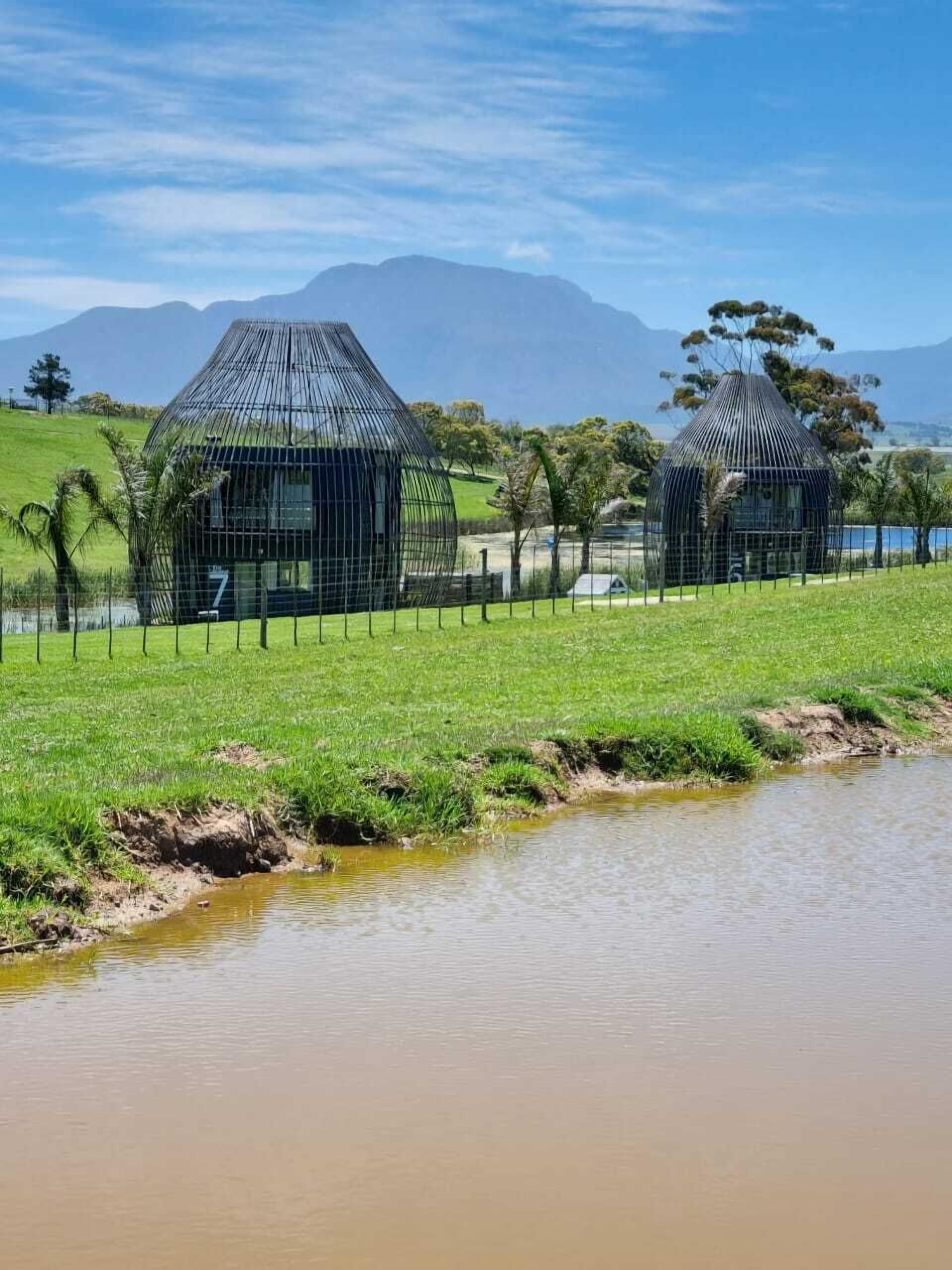 Dos estructuras modernas de forma abovedada con estructura metálica, rodeadas por un campo cubierto de hierba y una masa de agua, con montañas al fondo.