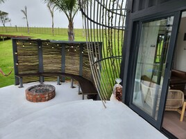 Un área de estar exterior moderna con un banco curvo, un pequeño hogar para fuego de ladrillo y una puerta de cristal que da a un espacio interior con una silla de mimbre.