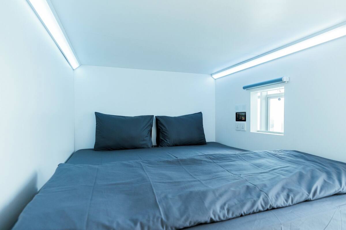 A small, modern bedroom with a single bed, two pillows, and a window with blinds.