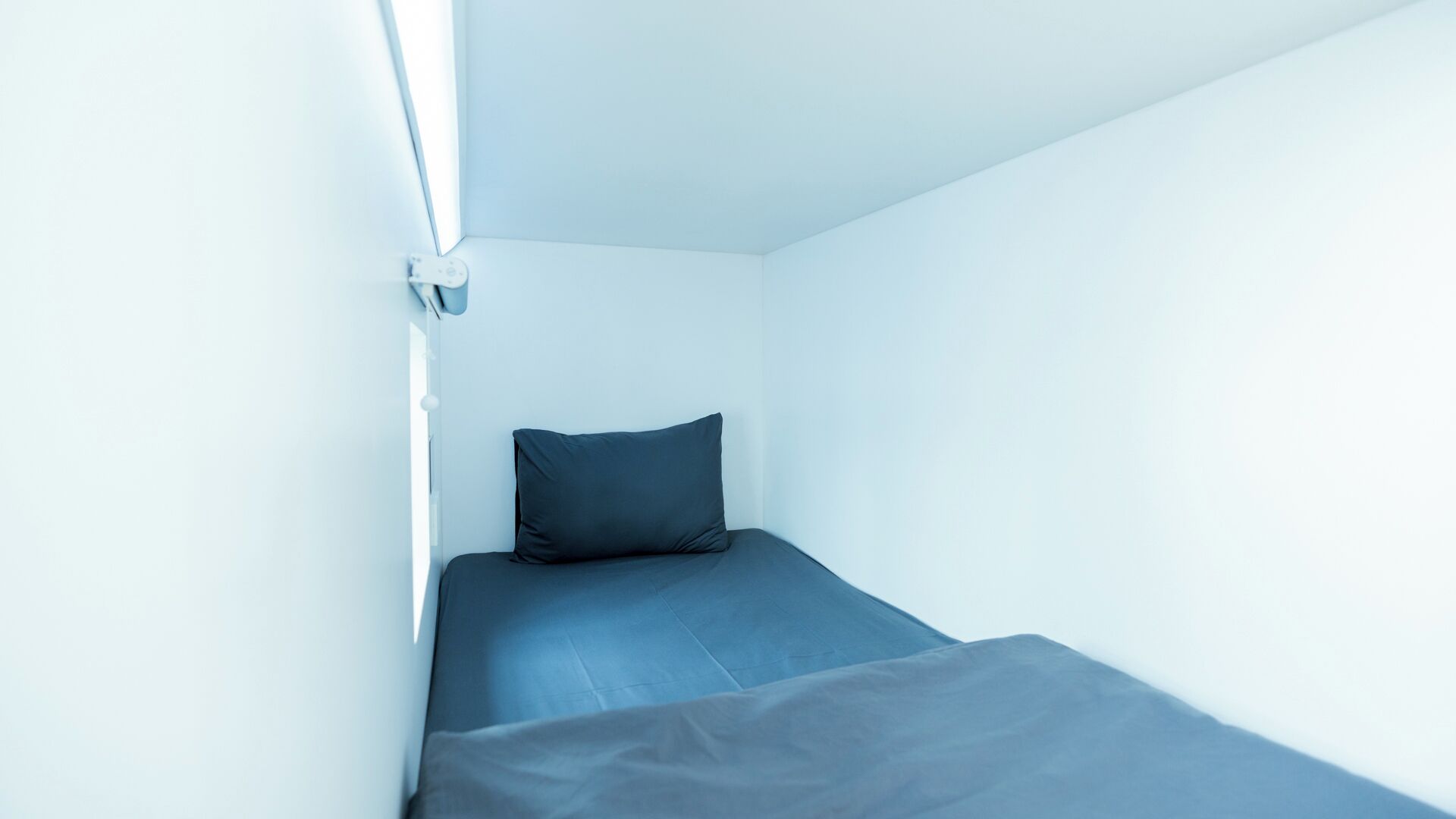 A small, single-bed room with a plain white wall and a single dark-colored pillow.