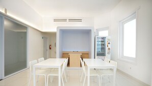 Una habitación moderna y luminosa con una mesa y sillas blancas, una pequeña cocina y un amplio ventanal que proporciona luz natural.
