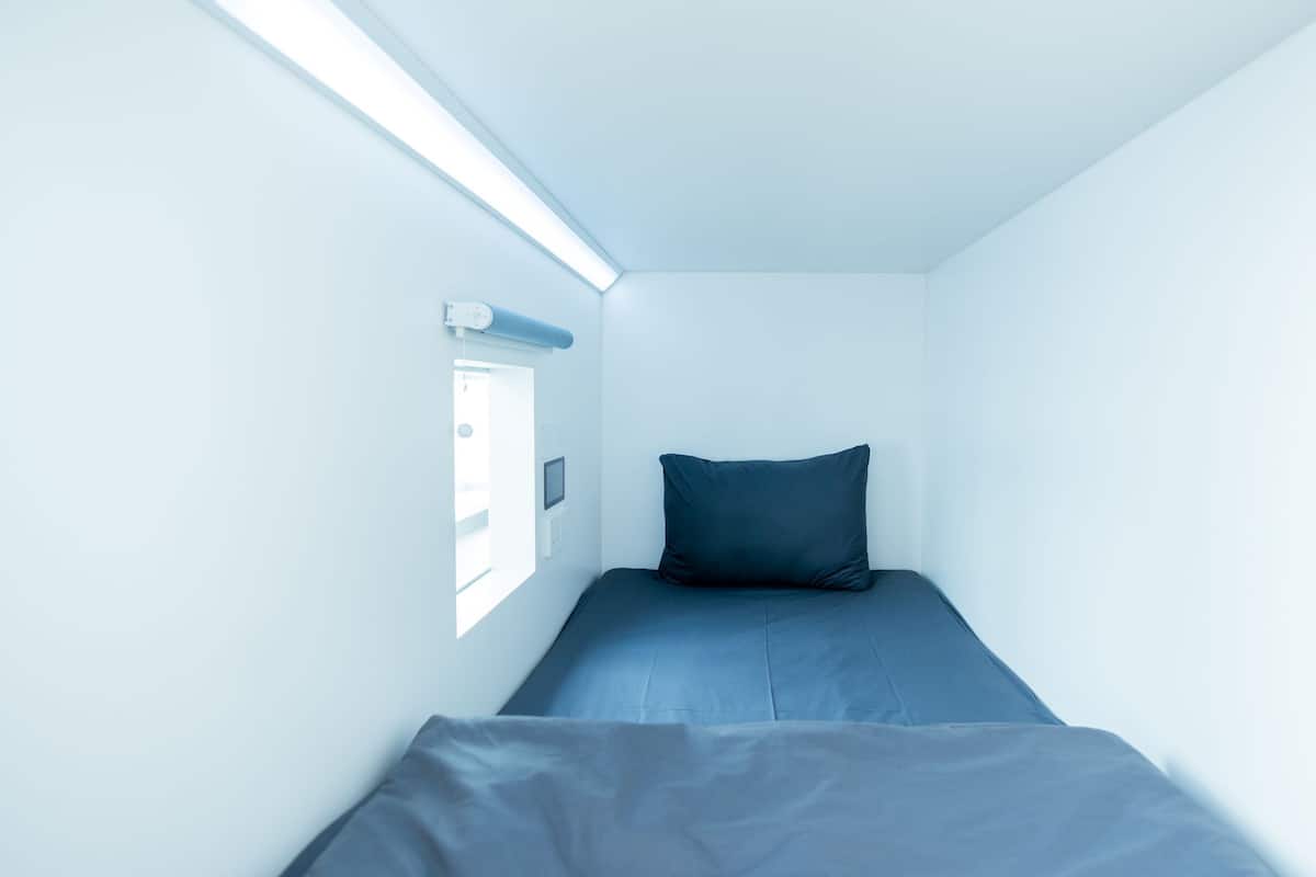 A small, narrow room with a single bed, a window, and a wall-mounted control panel.
