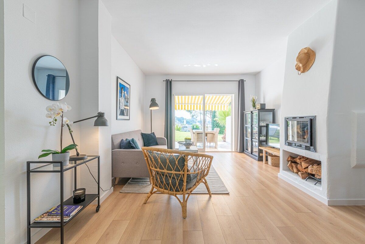 A modern living room with a sofa, a wicker chair, a small table with a lamp, a TV, and a fireplace.