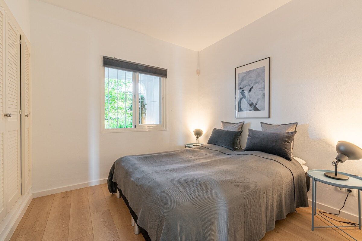 Ein Schlafzimmer mit einem Bett, zwei Nachttischen, einem Fenster mit Jalousien und einem gerahmten Bild an der Wand.