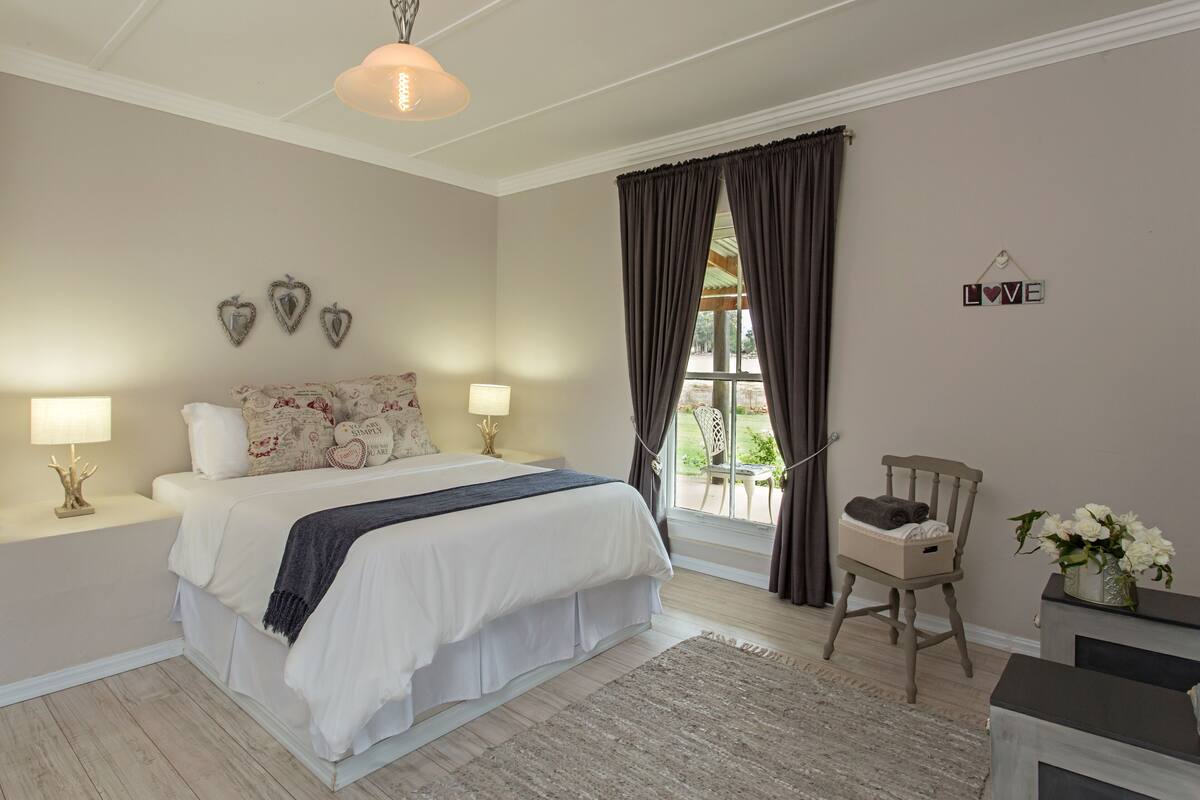 Een slaapkamer met een groot bed, twee bedlampen, een raam met gordijnen, een stoel met een mand, en een 'LOVE' bord op de muur.