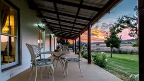 Alpendre coberto com mesa e cadeiras, com vista para uma paisagem gramada com árvores e um pôr do sol.