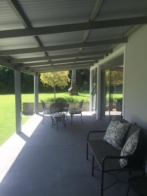 A covered patio area with a table and chairs, a cushioned bench, and a view of a well-maintained lawn and trees.