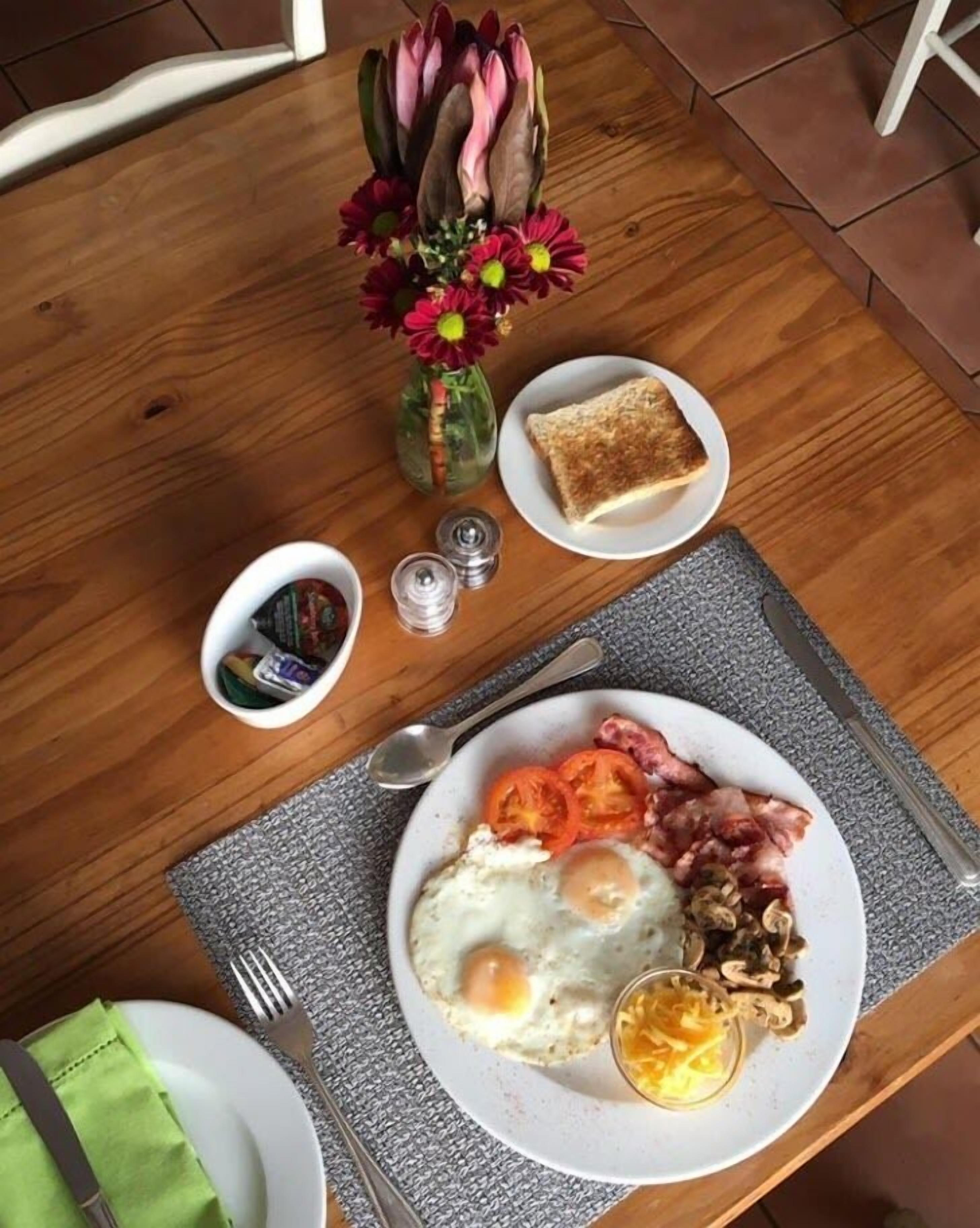 A breakfast setting with toast, eggs, tomatoes, and bacon on a plate, a vase of flowers, and condiments on a wooden table.