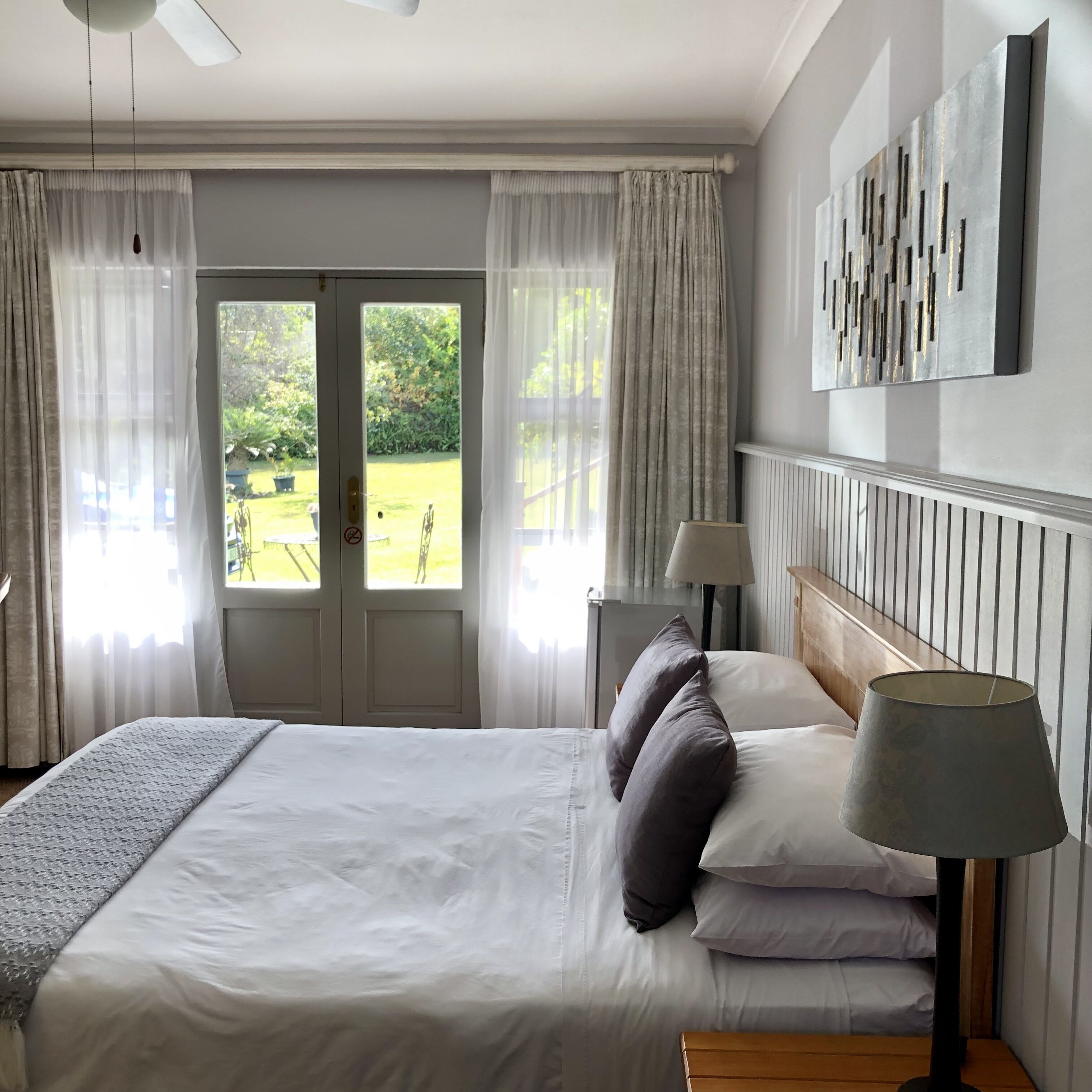 A hotel room with a bed, two bedside lamps, a view of the garden through a door, and a ceiling fan.