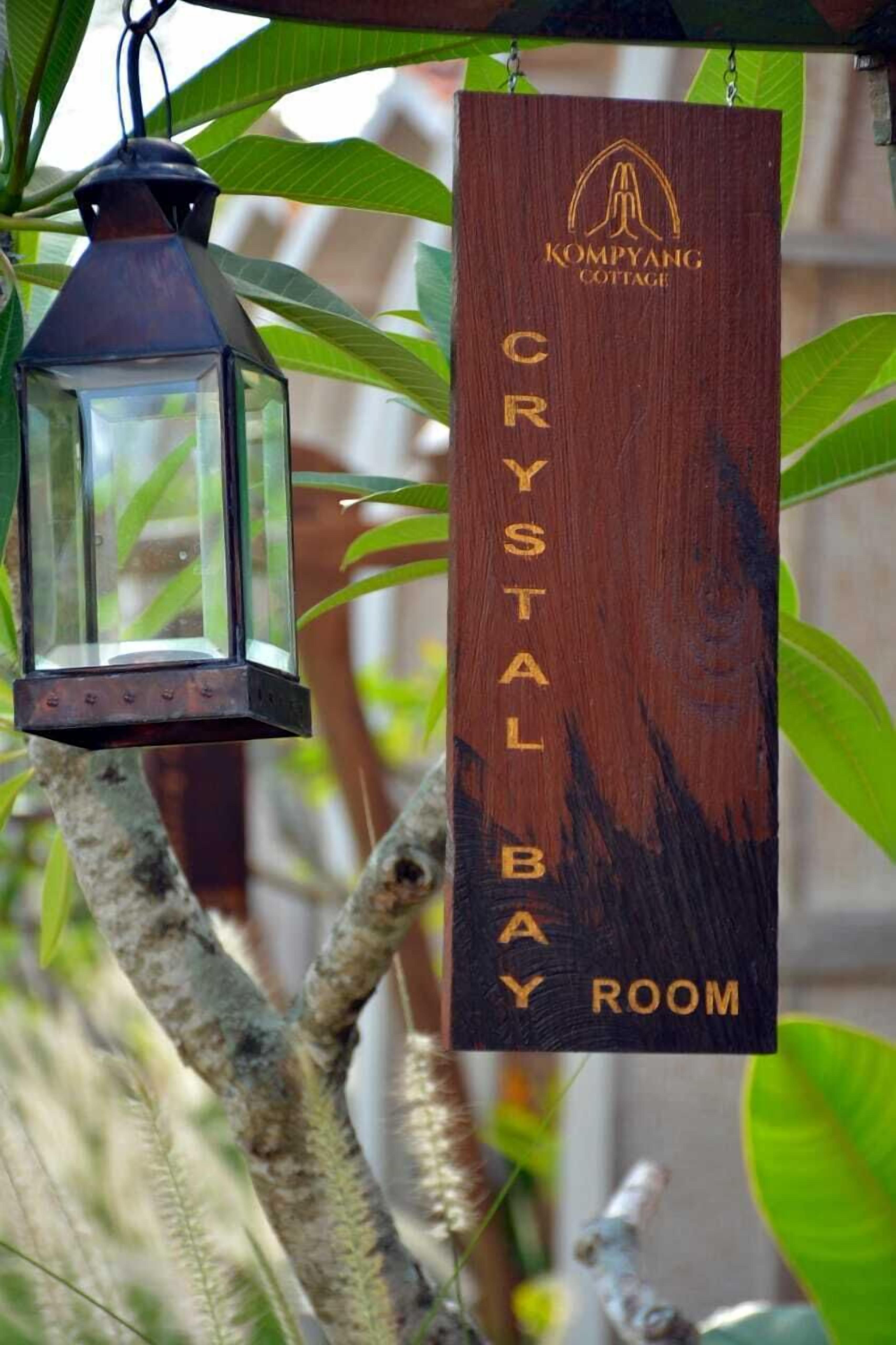 Et træskilt med teksten "KOMPYANG COTTAGE CRYSTAL BAAY ROOM" hænger i et træ.