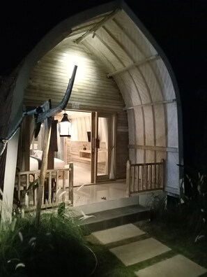 A cozy, semi-open arched structure with a wooden bench and a small table, leading to a warmly lit interior with a visible bathroom and a bed.