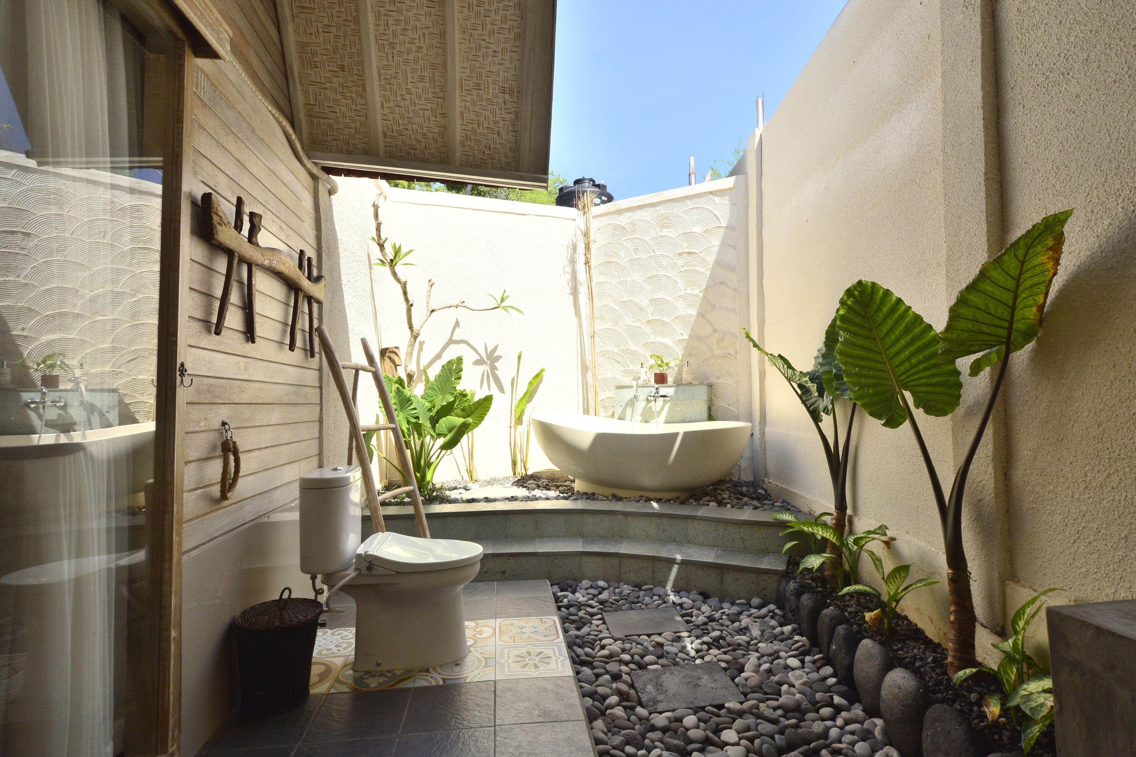 Un baño con bañera, inodoro y zona de ducha, rodeado de plantas y un sendero de guijarros.