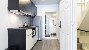 A narrow hallway with a kitchen area on the left, featuring a refrigerator, sink, and cabinets. A door leads to a staircase on the right. - Cozy Studio 1 in Sathorn - BTS Saint Louis (Bangkok)