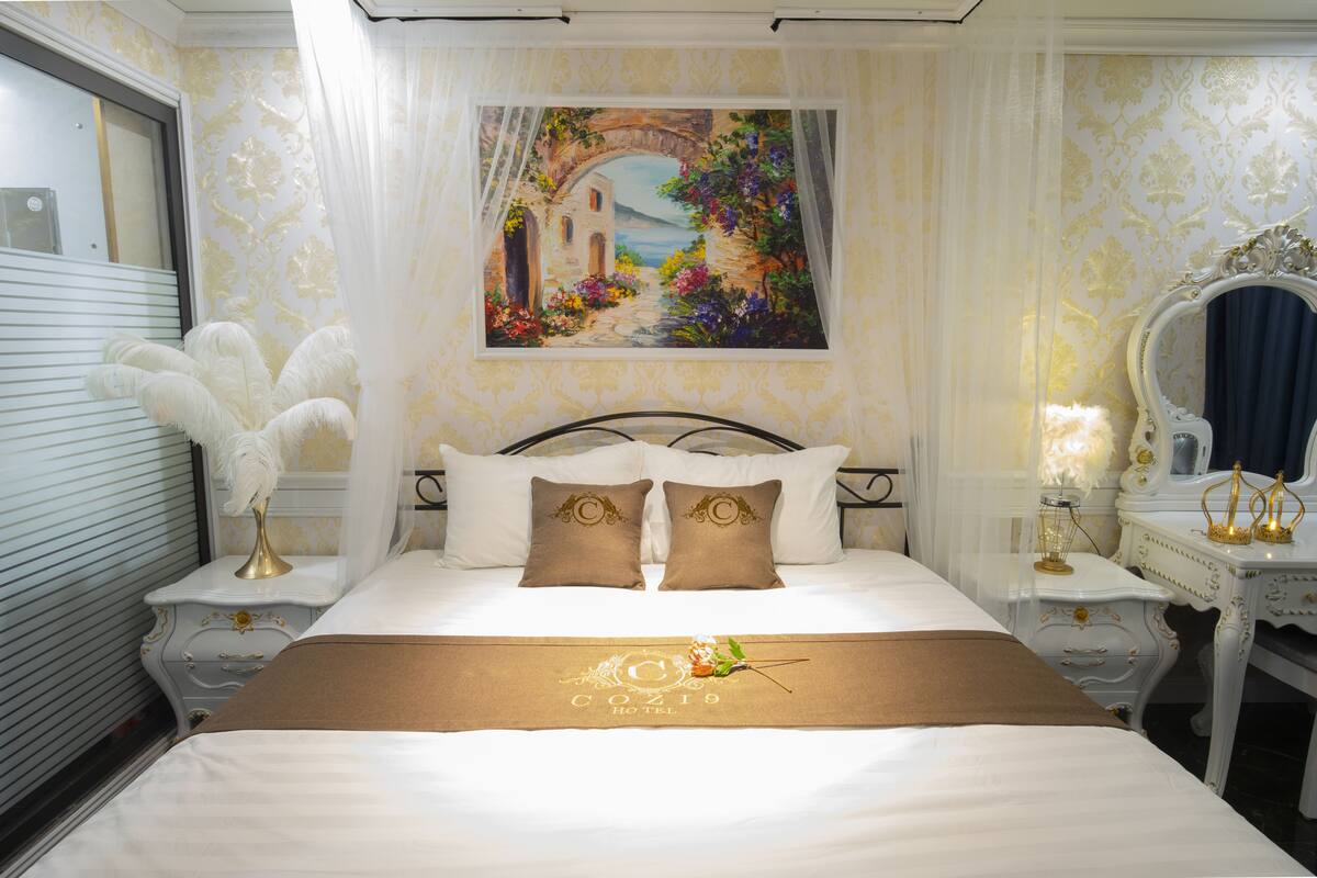 A hotel room with a bed, a painting of a garden, a mirror, and a decorative lamp.