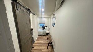 A hallway with a wooden floor, a sliding door, a bench, a bed with white bedding, and a window with blinds. - Historical “love” house downtown Kalispell Montana. (Kalispell)