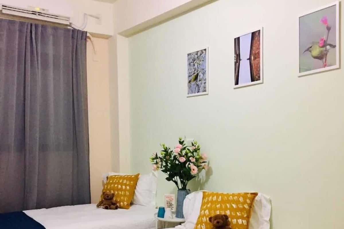 A hotel room with two beds, each with a blue blanket and a yellow pillow. There is a small table with a vase of flowers and a teddy bear on one bed. The walls are light green, and there are three framed pictures on the wall.
