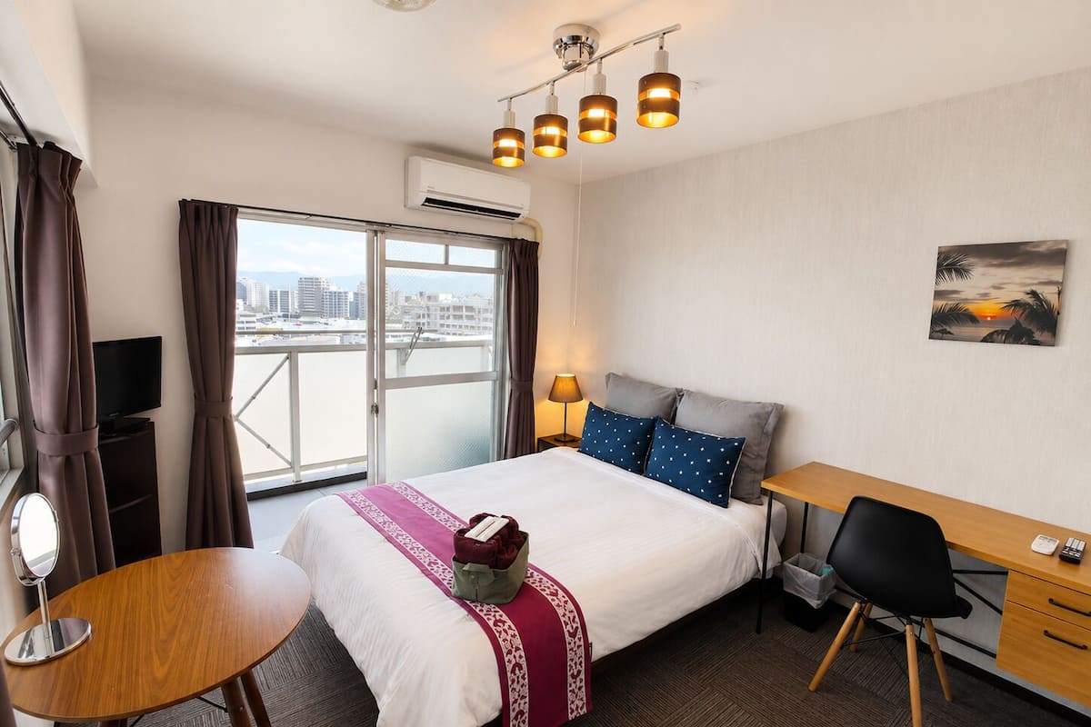 Habitación de hotel con cama, escritorio, silla y vista de la ciudad a través de un amplio ventanal.