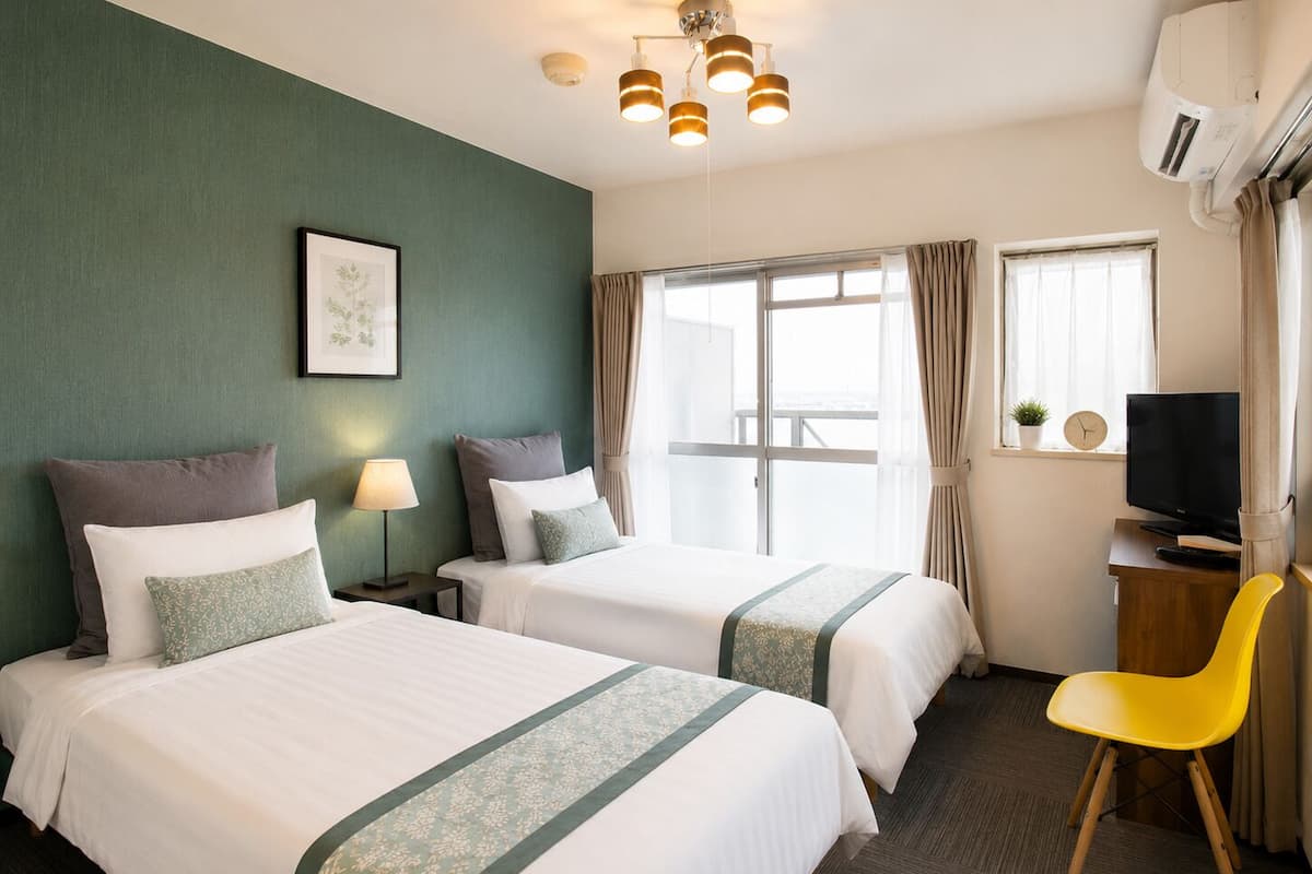 Habitación de hotel con dos camas, un escritorio, televisor, una silla y una ventana con cortinas.