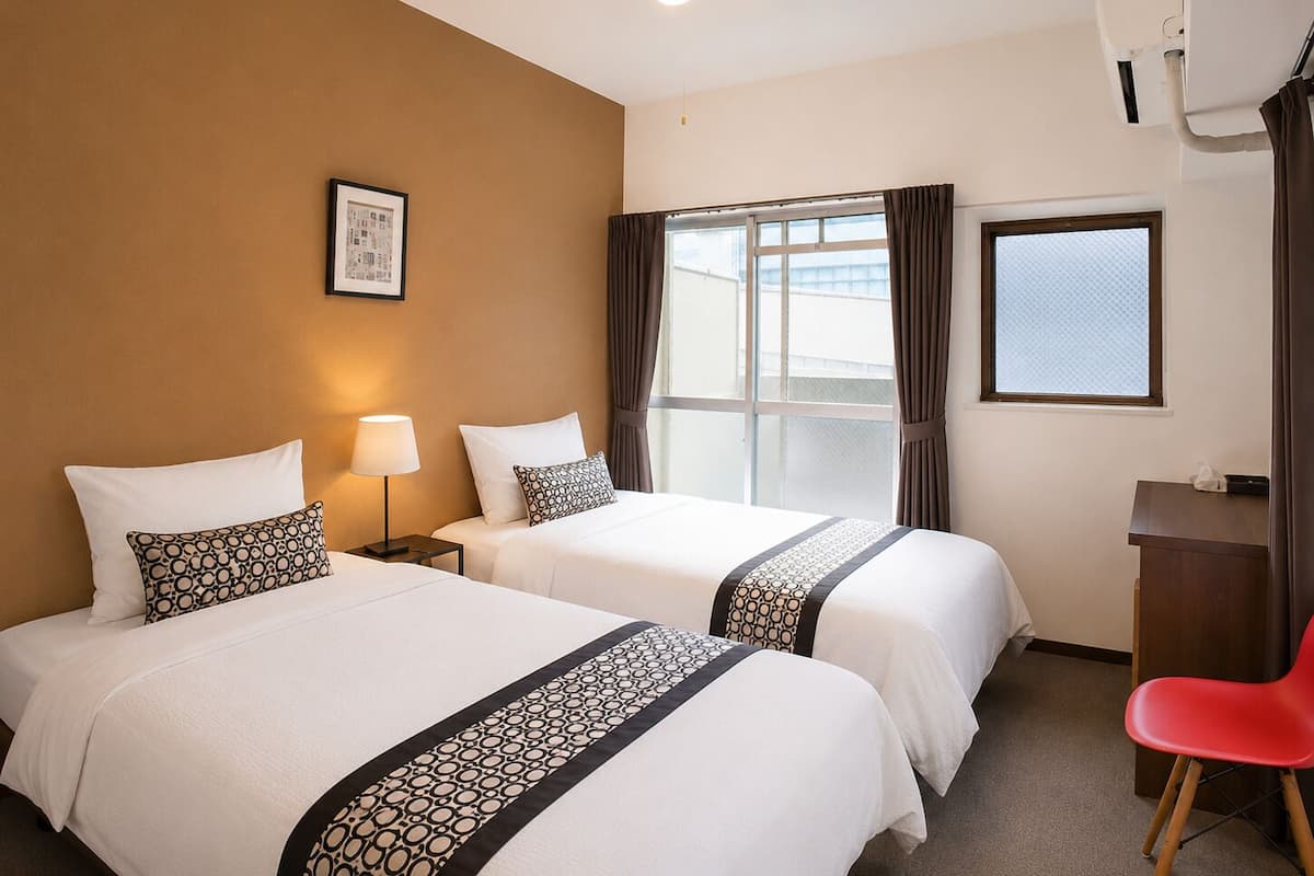 Habitación de hotel con dos camas, un escritorio, una silla, una ventana con cortinas, un cuadro enmarcado en la pared y un equipo de aire acondicionado.