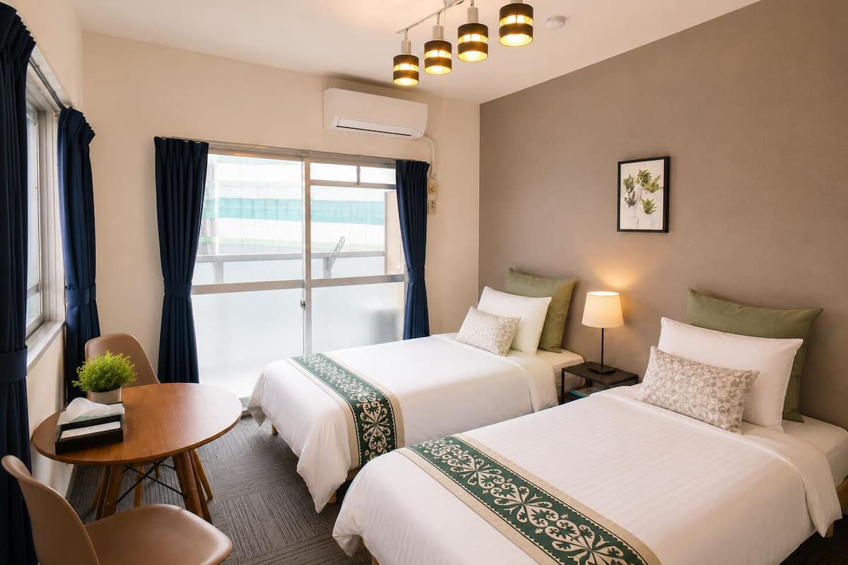Habitación de hotel con dos camas, un escritorio, una silla, ventana con cortinas, equipo de aire acondicionado y lámpara fijada a la pared.