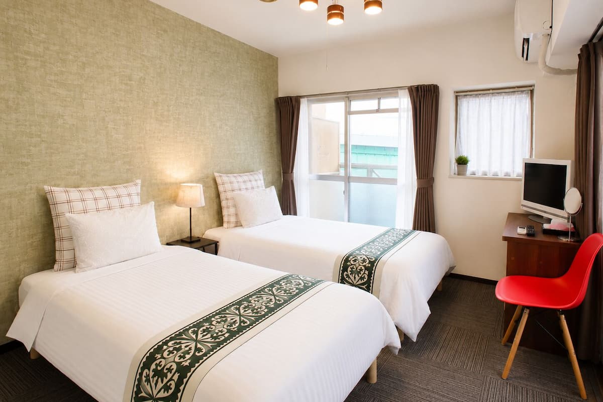 Habitación de hotel con dos camas, un escritorio con televisor, una silla roja y una ventana con cortinas.