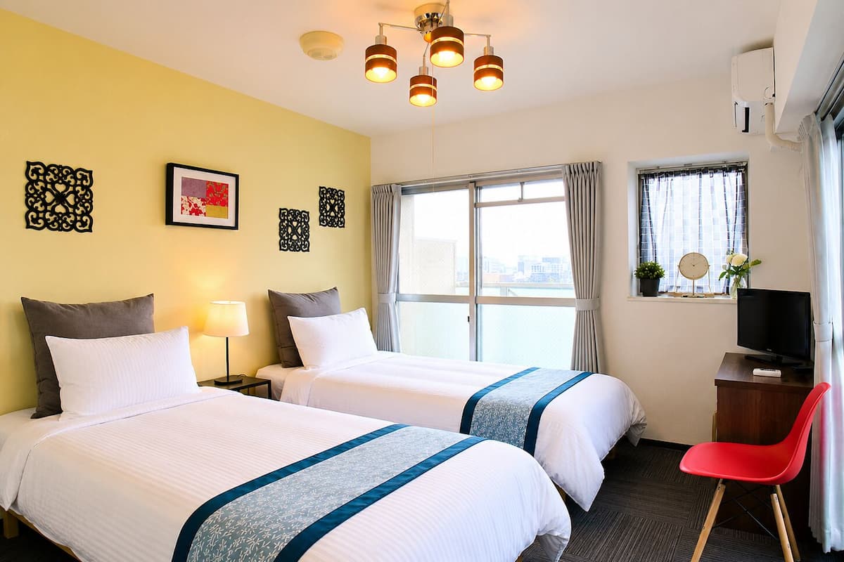 Habitación de hotel con dos camas, un escritorio con televisor, una silla roja y una ventana con vista a la ciudad.
