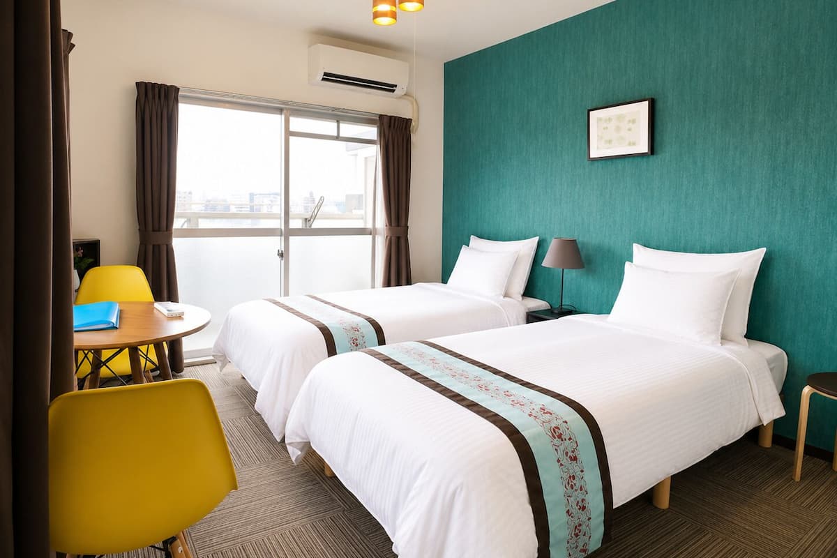 Habitación de hotel con dos camas, un escritorio, una silla, una lámpara y vista a la ciudad.
