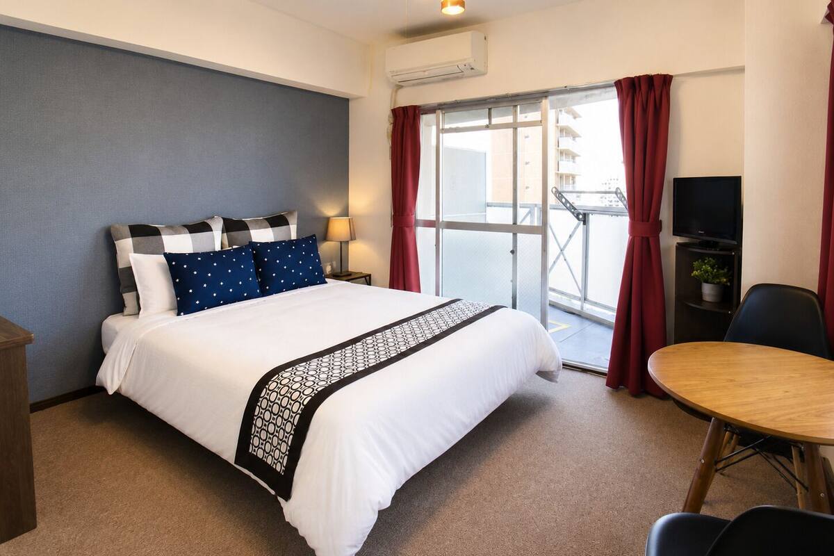 Habitación de hotel con cama, escritorio, silla, televisión y balcón con cortinas rojas.