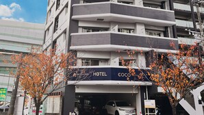 A multi-story hotel with balconies, a sign indicating 'HOTEL,' and a tree with autumn leaves in front. - COCO Fukuoka Chiyo (Fukuoka)
