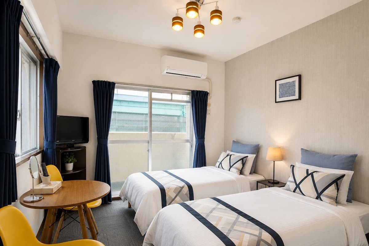 Habitación de hotel con dos camas, un televisor, una pequeña mesa con una silla y una ventana con cortinas.