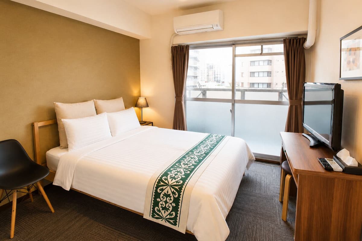 Habitación de hotel con cama, escritorio con televisor, equipo de aire acondicionado y vista a edificios a través de la ventana.