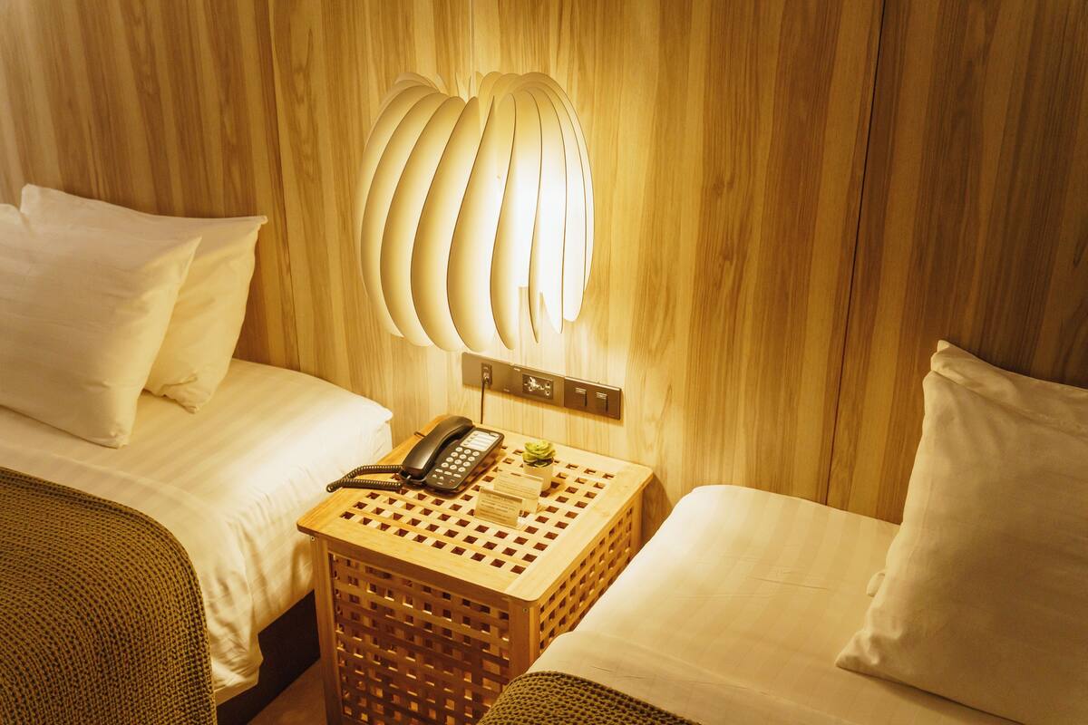 A hotel room with two beds, a wooden nightstand with a telephone and a small plant, and a unique wall-mounted light fixture.