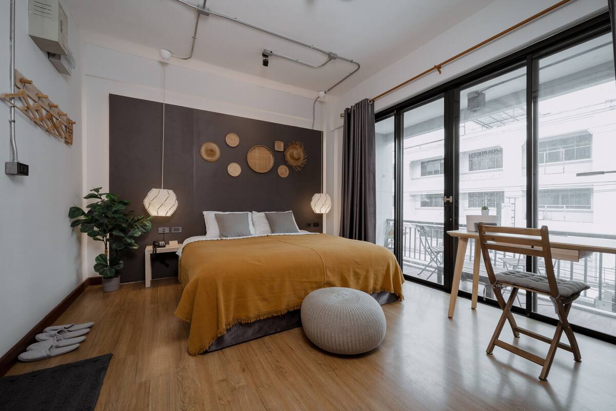 A modern bedroom with a large bed, a wooden floor, a balcony with a view, and a desk with a chair.