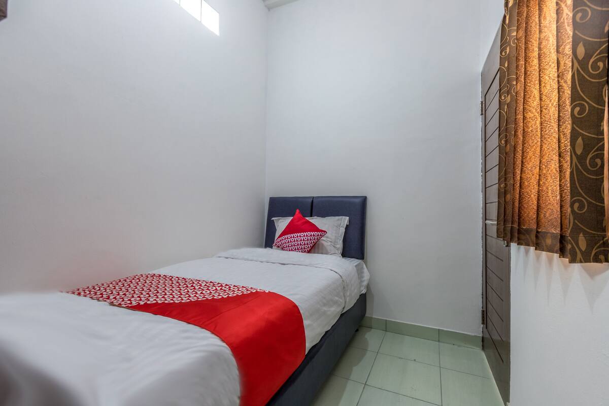 Une petite chambre avec un lit simple, une couvre-lit blanche et rouge, une tête de lit sombre et un rideau orné d’un motif de feuilles.