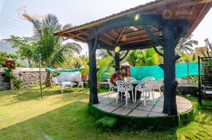 A covered outdoor seating area with a table and chairs, surrounded by palm trees and a white cow statue. - Malhar Villa (Murud)