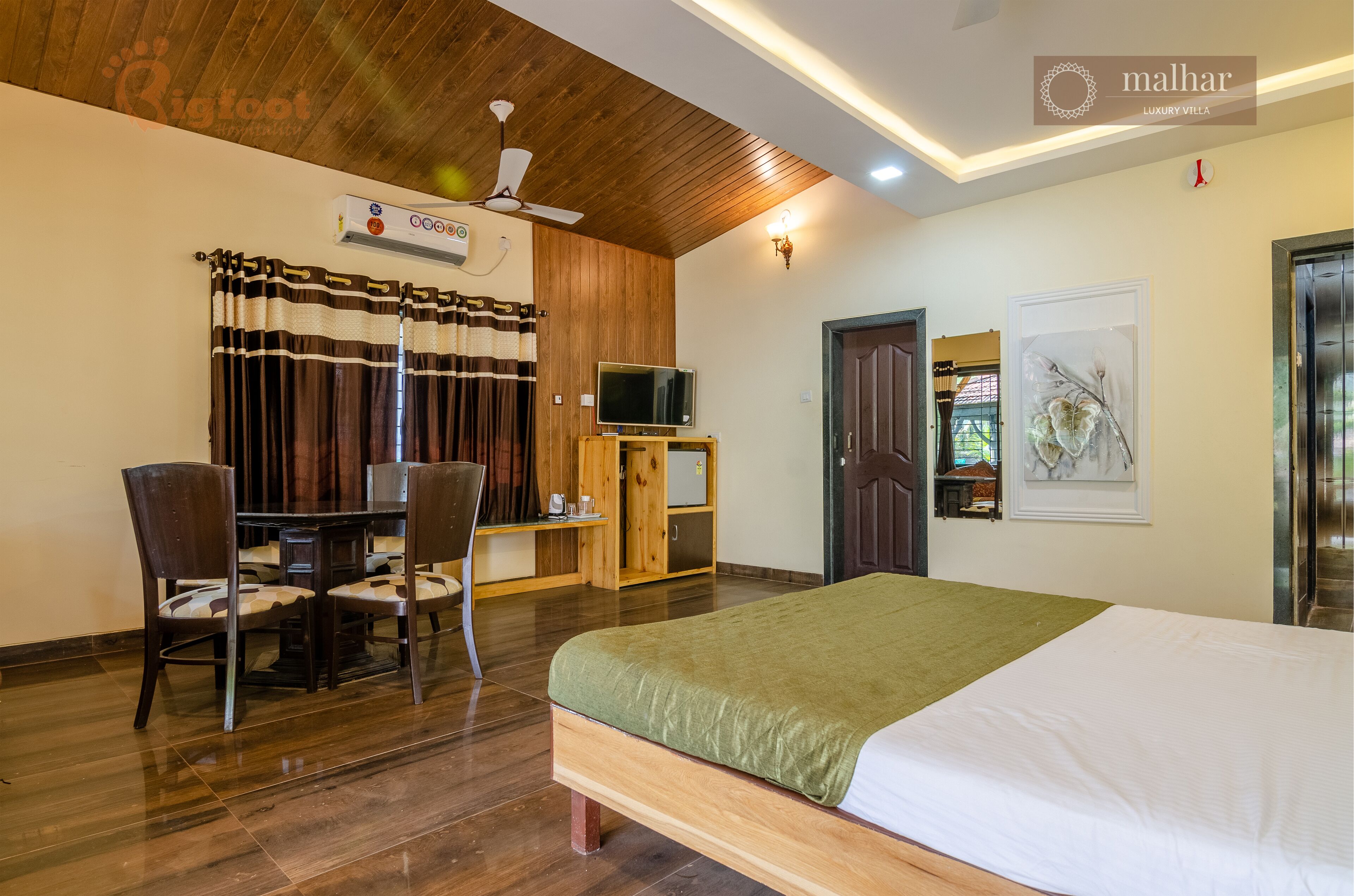 A hotel room with a wooden ceiling, a bed with a green bedspread, a desk with a television, and a chair.