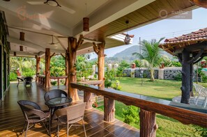 A covered outdoor area with wooden furniture and a view of a lush garden and mountains. - Malhar Villa (Murud)
