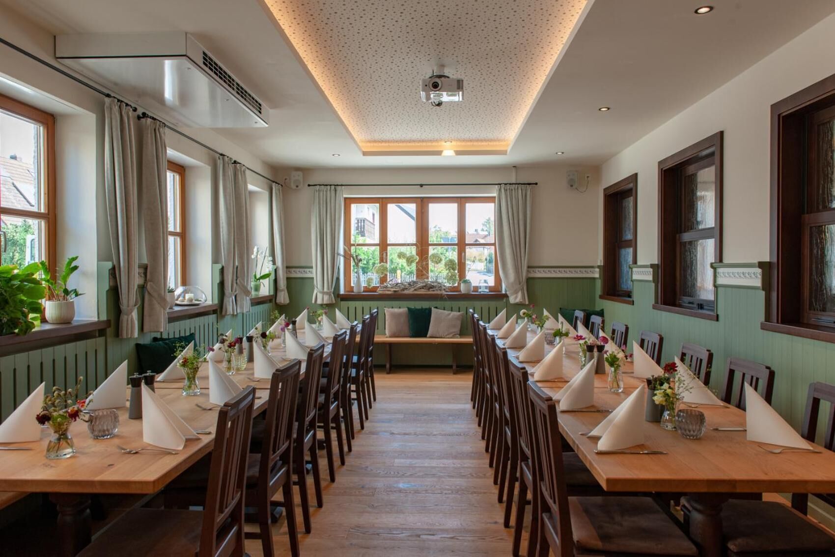 A long dining table set for a meal, with chairs on both sides. There are windows with curtains, a ceiling with a projector, and a bench with cushions.