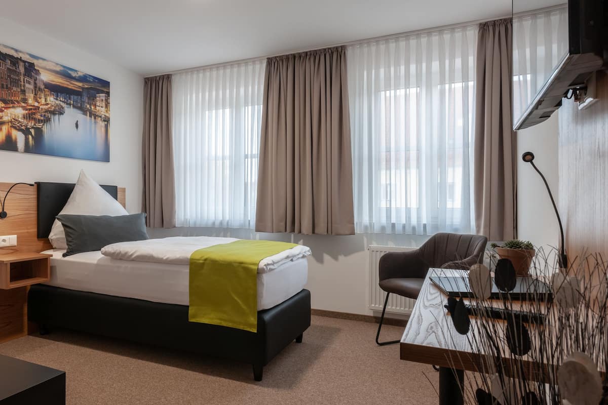 Ein modernes Hotelzimmer mit Bett, Schreibtisch, Stuhl, einem Fenster mit Vorhängen, einem Bild an der Wand und einer Heizung.