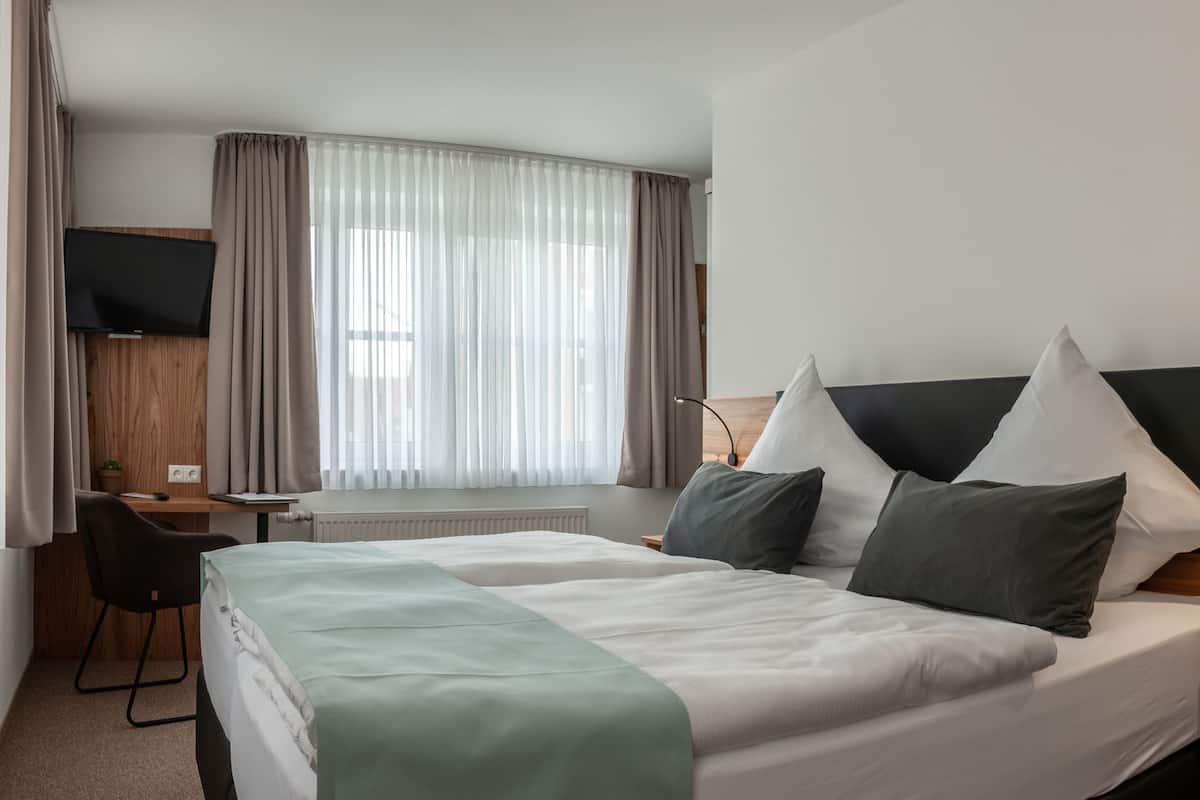 Ein Hotelzimmer mit Bett, Schreibtisch, Sessel, Fernseher und einem Fenster mit Vorhängen.