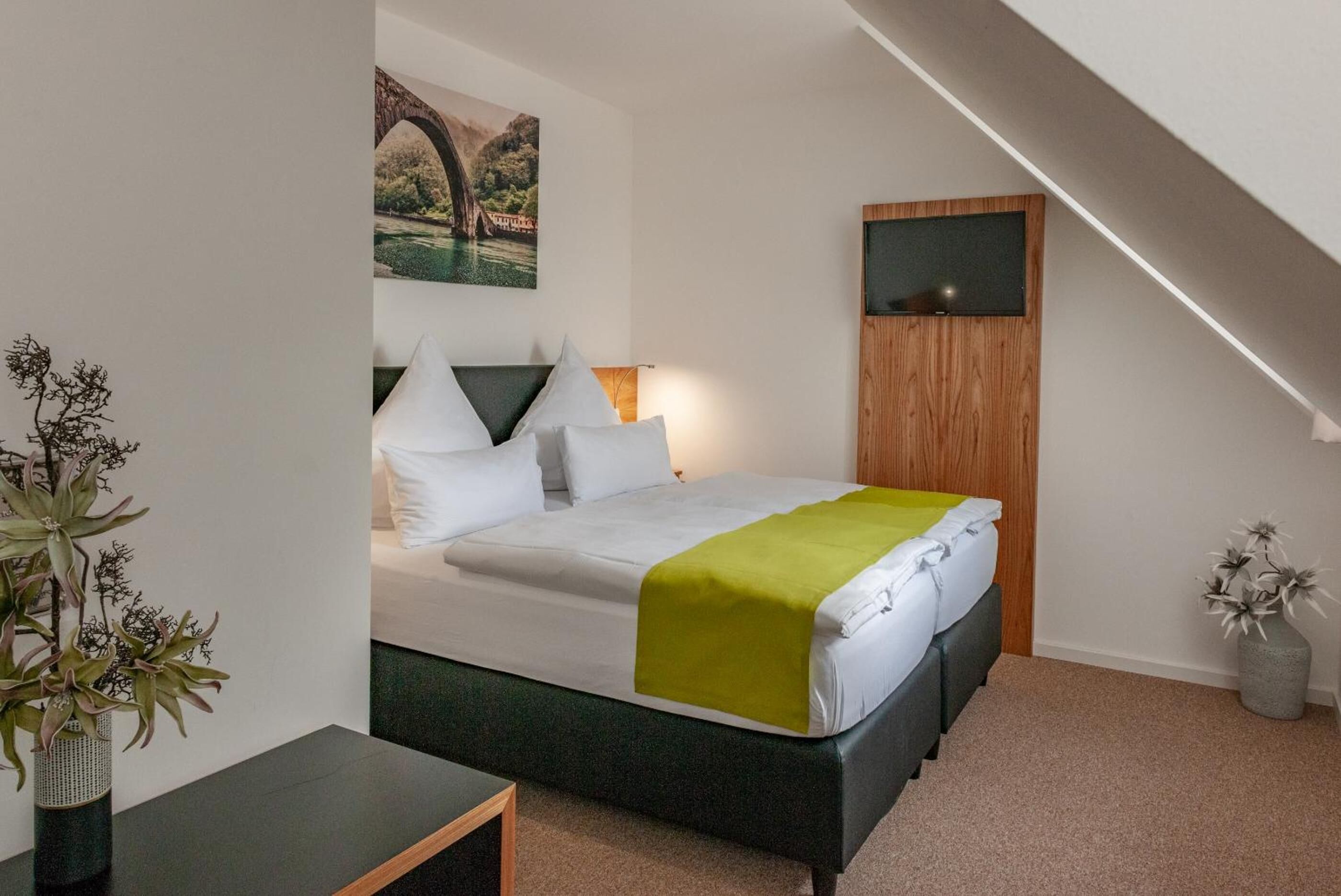 A bedroom with a bed, a painting of a bridge, a TV, and a plant.