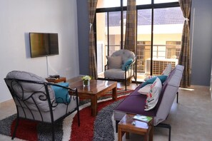 Una sala de estar con sofá, sillón y mesa de centro, un televisor de pantalla plana montado en la pared y vistas al balcón exterior a través de amplias ventanas.