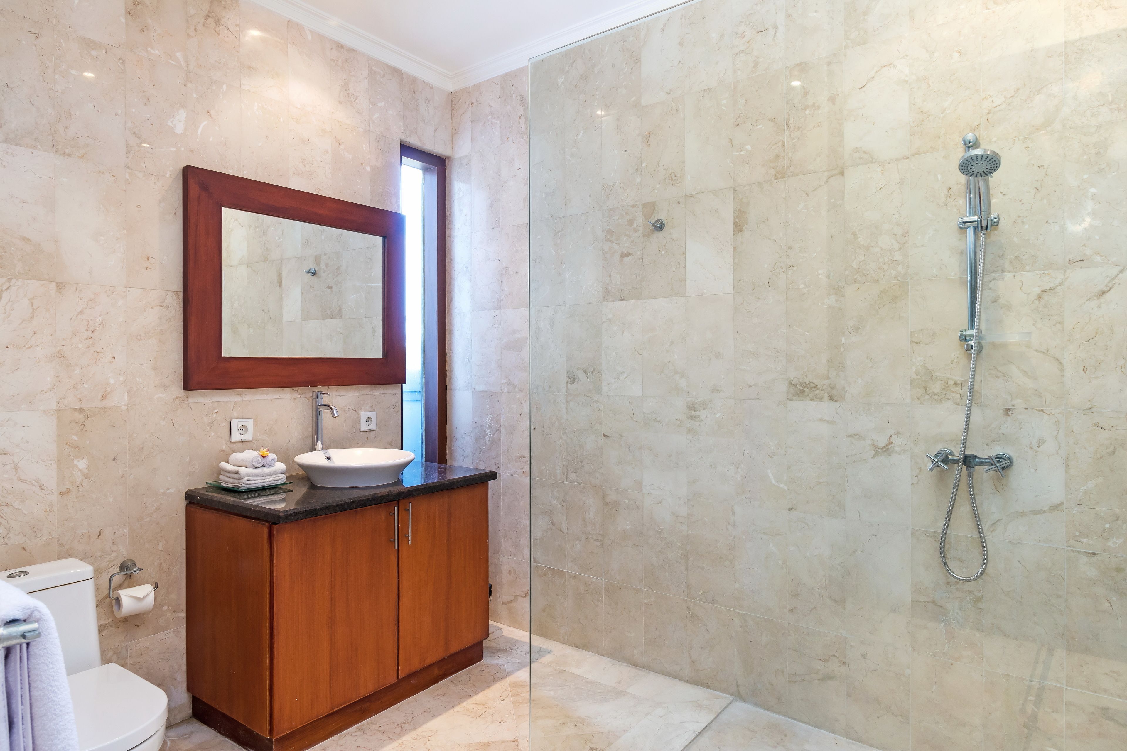 Un baño con pared de mármol, un mueble de madera, un lavabo, un inodoro y una ducha.