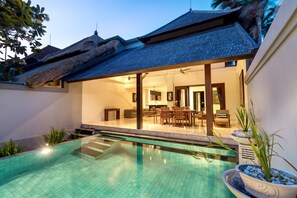 A modern pool area with a clear pool, a dining area with chairs and a table, and a thatched roof structure. - Villa Lalang Temu (Jimbaran)