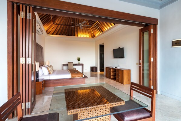 A modern bedroom with a glass coffee table, wooden furniture, a ceiling fan, and a TV on the wall. - Villa Lalang Temu (Jimbaran)